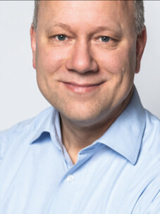 Porträt von Bert Hecht mit blauem Hemd vor hellem Hintergrund.